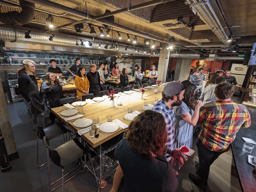 A group of people stand surrounding a dinner table.