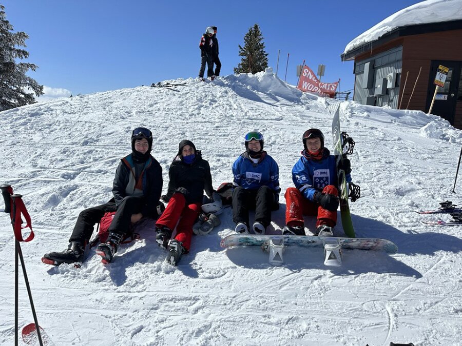 A group of people sit on a snowy hill in snowboarding attire.