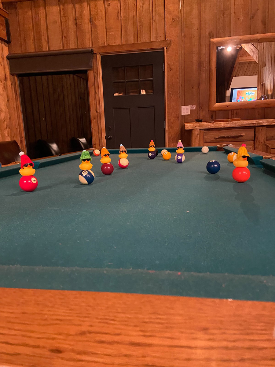 Several rubber duck toys in winter hats and sunglasses placed on top of pool balls on a pool table.